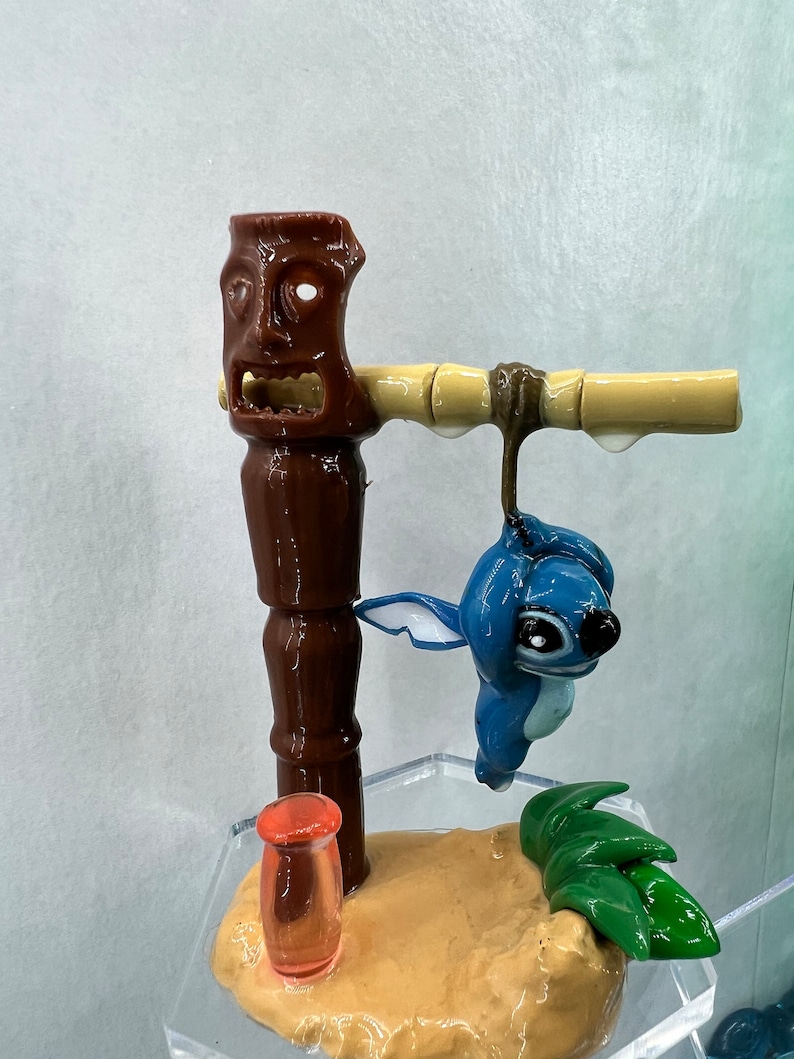 Pu&ograve; includere: Una statuina blu del personaggio Stitch appesa a un totem tiki marrone con un palo di bamb&ugrave;. La statuina &egrave; sospesa sopra una base sabbiosa con foglie verdi e un tubo di vetro rosso trasparente.