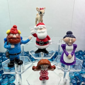 May include: A set of six Christmas-themed figurines, including Santa Claus, a reindeer, a woman in a purple dress, a man with a beard and a blue jacket, and two children. The figurines are displayed on a tiered hexagonal stand.