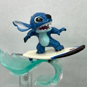 Pu&ograve; includere: Figurina blu di Stitch che cavalca un surf bianco con una striscia nera e rossa su un'onda blu.