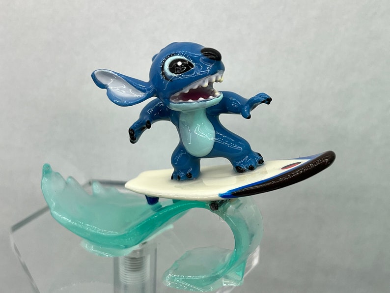 Pu&ograve; includere: Figurina blu di Stitch che cavalca un surf bianco con una striscia nera e rossa su un'onda blu.