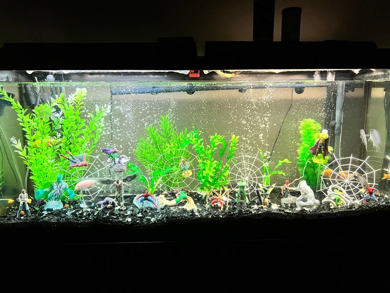 May include: A decorated aquarium with green plants, miniature superhero figurines, and white spiderwebs. The tank is filled with water and has a black gravel base. The aquarium is lit from above.