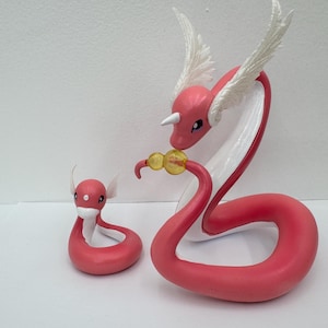 Pode incluir: Duas figuras rosa e brancas de uma criatura serpentina com asas brancas e um chifre. Uma é maior, com um orbe amarelo, e a outra é menor. As figuras estão em uma superfície branca.
