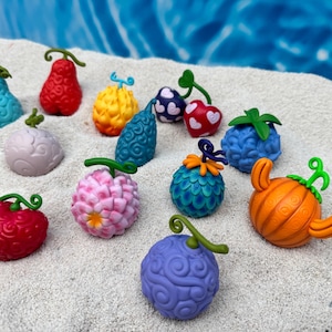 Puede incluir: Colección de figuras de frutas coloridas y estilizadas en varias formas y colores, incluyendo azul, rojo, amarillo y morado. Cada fruta tiene un tallo verde y se encuentra sobre una superficie arenosa.