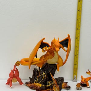 May include: A collection of orange and black Charizard figurines, with flames and a rocky base. The figures are on a white surface. A yellow ruler is in the background.