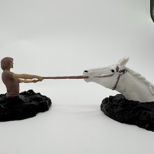 Peut inclure: Un ensemble de figurines représentant une personne en t-shirt marron et une tête de cheval blanc, émergeant toutes deux de bases noires texturées distinctes. La personne tient une fine corde marron reliée à la bouche du cheval.