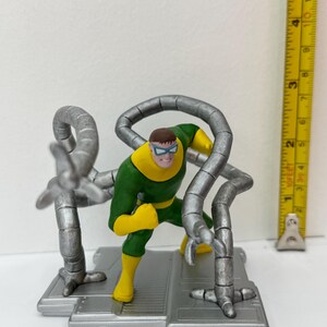 May include: A Dr. Octopus action figure. The figure is in a dynamic pose, wearing a green and yellow suit with silver mechanical arms. The figure is on a gray platform. A ruler is next to the figure.
