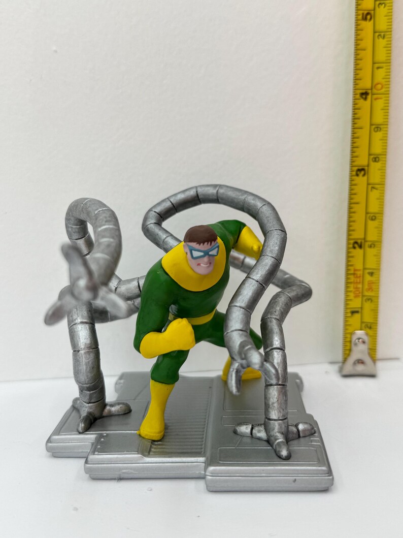 May include: A Dr. Octopus action figure. The figure is in a dynamic pose, wearing a green and yellow suit with silver mechanical arms. The figure is on a gray platform. A ruler is next to the figure.