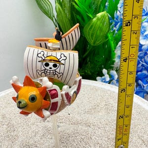 Puede incluir: Figura detallada de un barco de dibujos animados con una cara de sol. El barco tiene un casco blanco con rayas negras y un emblema de Jolly Roger. La figura mide aproximadamente 5 cm de altura.