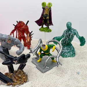 May include: A collection of superhero action figures, including Rhino, Venom, and Doctor Octopus. The figures are brightly colored and detailed, with a variety of poses and designs. The figures are on a sandy surface.