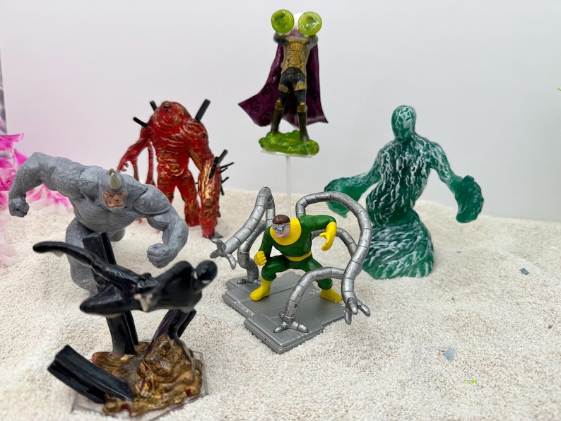 May include: A collection of superhero action figures, including Rhino, Venom, and Doctor Octopus. The figures are brightly colored and detailed, with a variety of poses and designs. The figures are on a sandy surface.