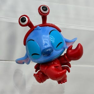 May include: A blue and red Stitch figurine wearing a red crab costume with large, white eyes.