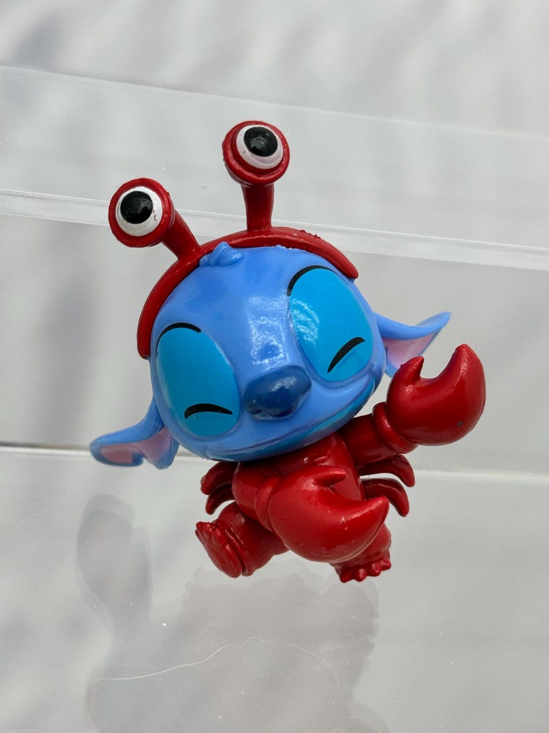 May include: A blue and red Stitch figurine wearing a red crab costume with large, white eyes.