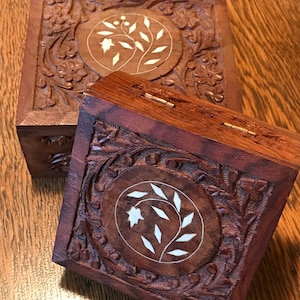 May include: Two intricately carved wooden boxes with floral designs. The boxes are made of dark wood with white inlay. The top box is open, revealing the smaller box inside.