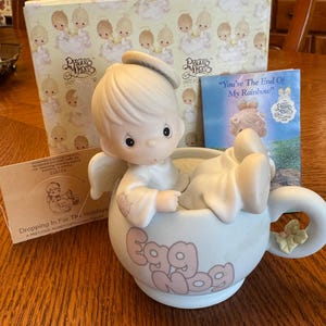 May include: A Precious Moments figurine of an angel in a teacup that says "Egg Nog". The figurine is light-colored with a halo and wings. The box and a small card are in the background.