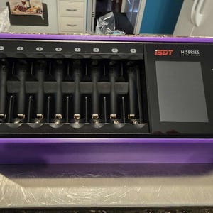 May include: A purple ISDT N Series smart charger with eight charging slots. The charger has a black screen and the ISDT logo is visible. The charger is on a metal surface.