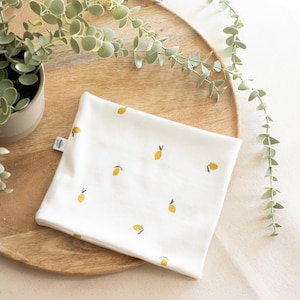 May include: A white baby bib with a pattern of yellow lemons. The bib is folded and placed on a wooden tray, with greenery in the background. The bib has a small tag on the side.