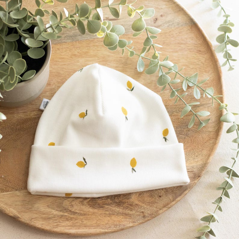 May include: A white ribbed knit beanie featuring a pattern of yellow lemons. The hat has a folded brim and is likely made of soft, comfortable material. The beanie is a stylish accessory for cooler weather.