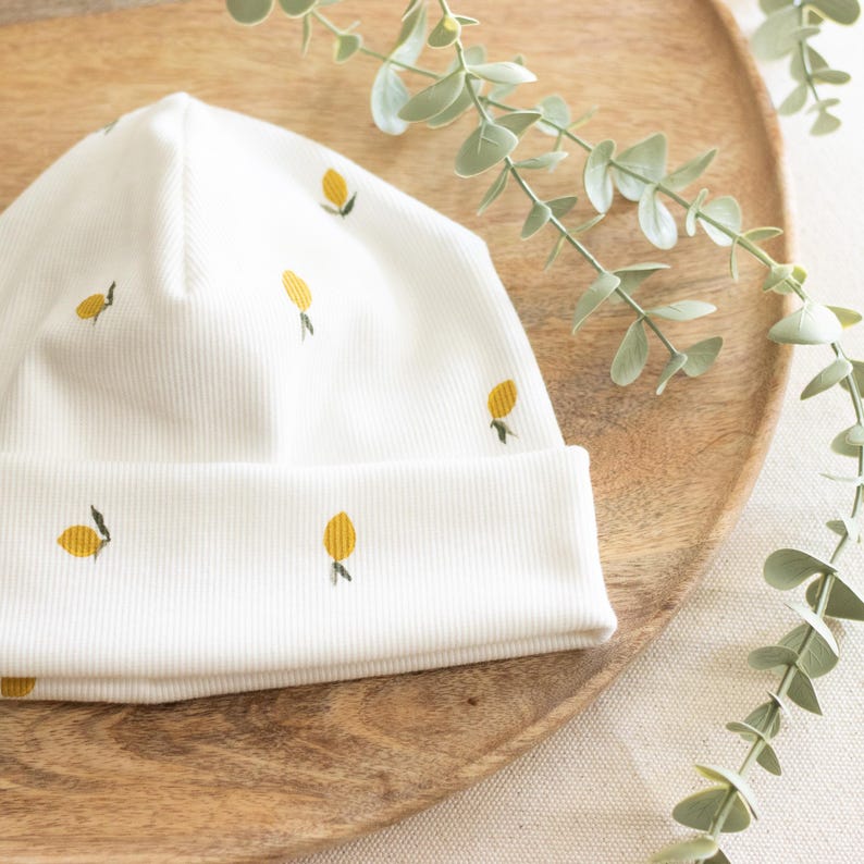 May include: A white ribbed knit beanie featuring embroidered yellow lemons. The hat is folded at the brim and sits on a wooden tray with a sprig of greenery. The design is simple and fresh.