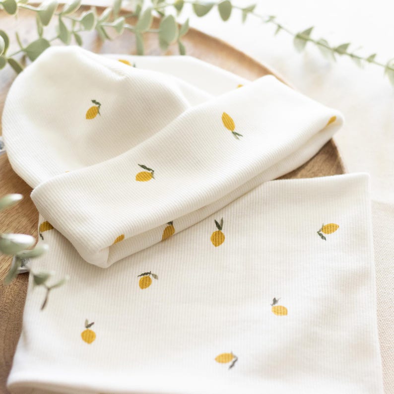 May include: Cream-colored baby hat and neck warmer set. The ribbed fabric is adorned with a repeating pattern of small yellow lemons with green leaves. The set is displayed on a wooden tray, suggesting a natural aesthetic.