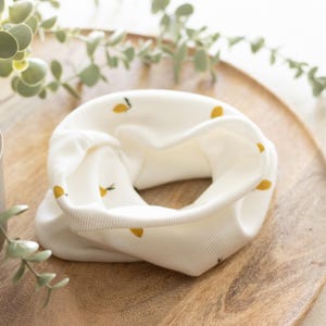 May include: A cream-colored neck gaiter with a repeating pattern of small yellow lemons. The fabric is ribbed and appears soft. The gaiter is folded in a circular shape, showcasing the interior and exterior. The background is a wooden tray.