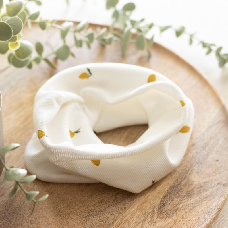 May include: A cream-colored neck gaiter with a repeating pattern of small yellow lemons. The fabric is ribbed and appears soft. The gaiter is folded in a circular shape, showcasing the interior and exterior. The background is a wooden tray.