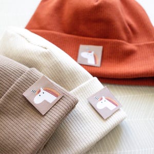 Könnte beinhalten: Drei Strickmützen in verschiedenen Farben: Braun, Beige und Orange. Jede Mütze hat einen kleinen gestickten Einhorn-Aufnäher.