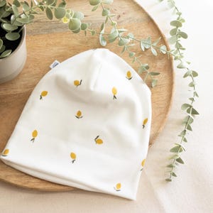 May include: A white beanie with a ribbed texture, adorned with small yellow floral embroidery. The hat is made of a soft material and is suitable for babies or children. The beanie is displayed on a wooden tray with greenery.