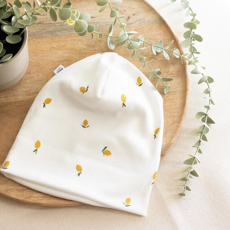 May include: A white beanie with a ribbed texture, adorned with small yellow floral embroidery. The hat is made of a soft material and is suitable for babies or children. The beanie is displayed on a wooden tray with greenery.