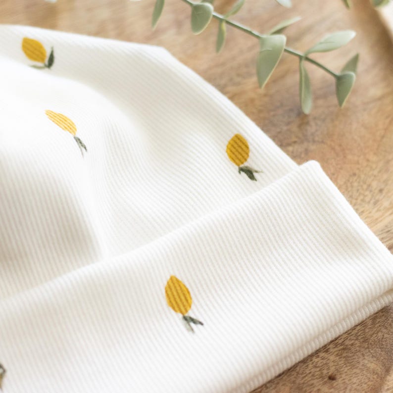 May include: A white ribbed knit beanie featuring a repeating pattern of yellow lemons with green leaves. The hat is folded at the brim and is made of a soft, comfortable material. The design is simple and cheerful.