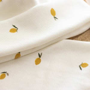 May include: Close-up of white fabric with a ribbed texture, adorned with small embroidered yellow lemons and green leaves. The fabric is folded, revealing multiple layers. The overall theme is a fresh, summery design.