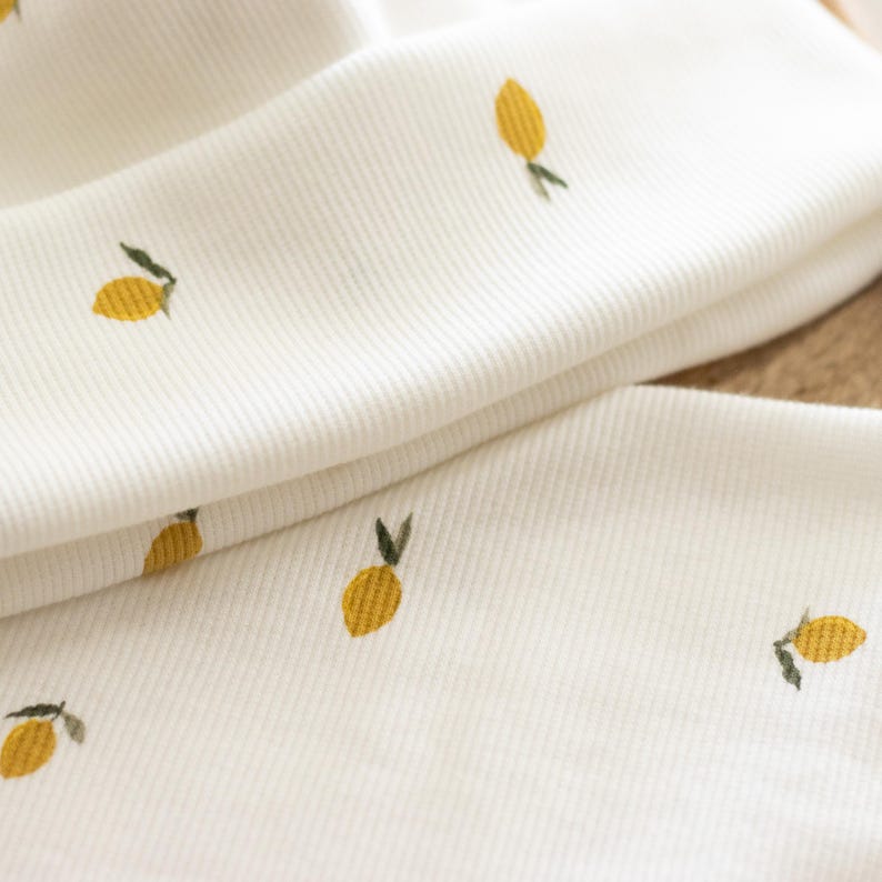 May include: Close-up of white fabric with a ribbed texture, adorned with small embroidered yellow lemons and green leaves. The fabric is folded, revealing multiple layers. The overall theme is a fresh, summery design.