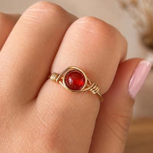 May include: A gold-toned wire-wrapped ring featuring a single, round, deep red gemstone at its centre. The metal wire is coiled around the base of the stone, creating a circular frame that connects to a thin, smooth band encircling the finger.