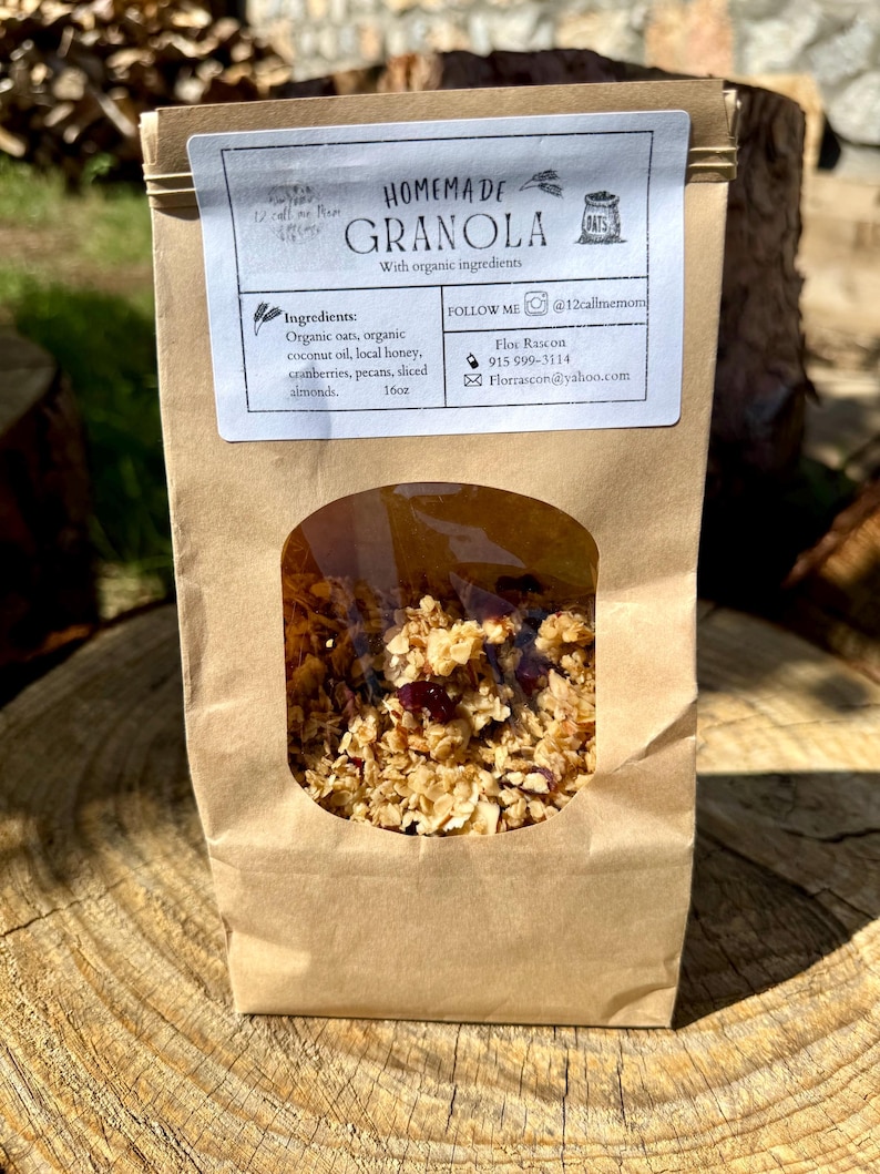 May include: A brown paper bag of homemade granola with a clear window, sitting on a tree stump. The label reads "Homemade Granola" and lists ingredients like organic oats, coconut oil, and cranberries. The bag contains 16oz of granola.