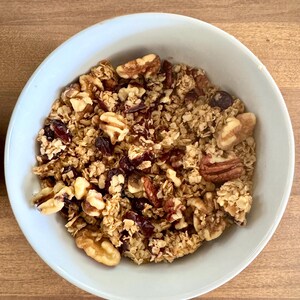 May include: A light blue bowl filled with granola. The granola contains oats, walnuts, pecans, and dried cranberries. The bowl is sitting on a wooden surface.