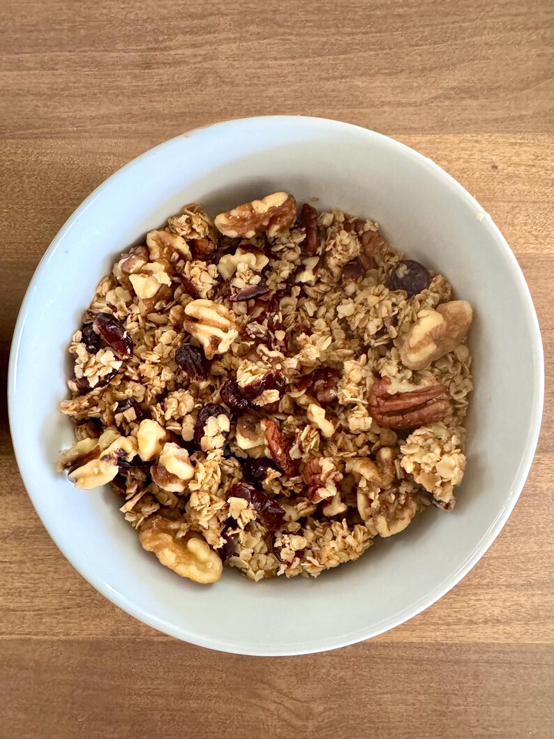 May include: A light blue bowl filled with granola. The granola contains oats, walnuts, pecans, and dried cranberries. The bowl is sitting on a wooden surface.