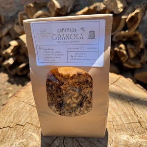 May include: A brown paper bag of homemade granola with a clear window, displaying the contents. The label reads "Homemade Granola" with a list of ingredients: organic oats, coconut oil, local honey, cranberries, walnuts, and pecans. The bag is on a wooden surface.