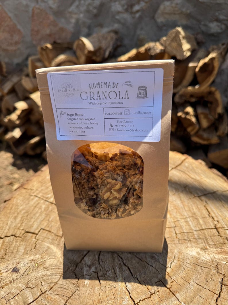 May include: A brown paper bag of homemade granola with a clear window, displaying the contents. The label reads "Homemade Granola" with a list of ingredients: organic oats, coconut oil, local honey, cranberries, walnuts, and pecans. The bag is on a wooden surface.
