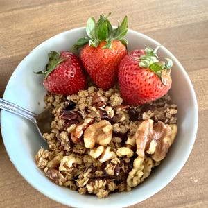 May include: A light blue bowl filled with granola, walnuts, and three fresh strawberries. A silver spoon rests in the bowl. The food is on a wooden table.
