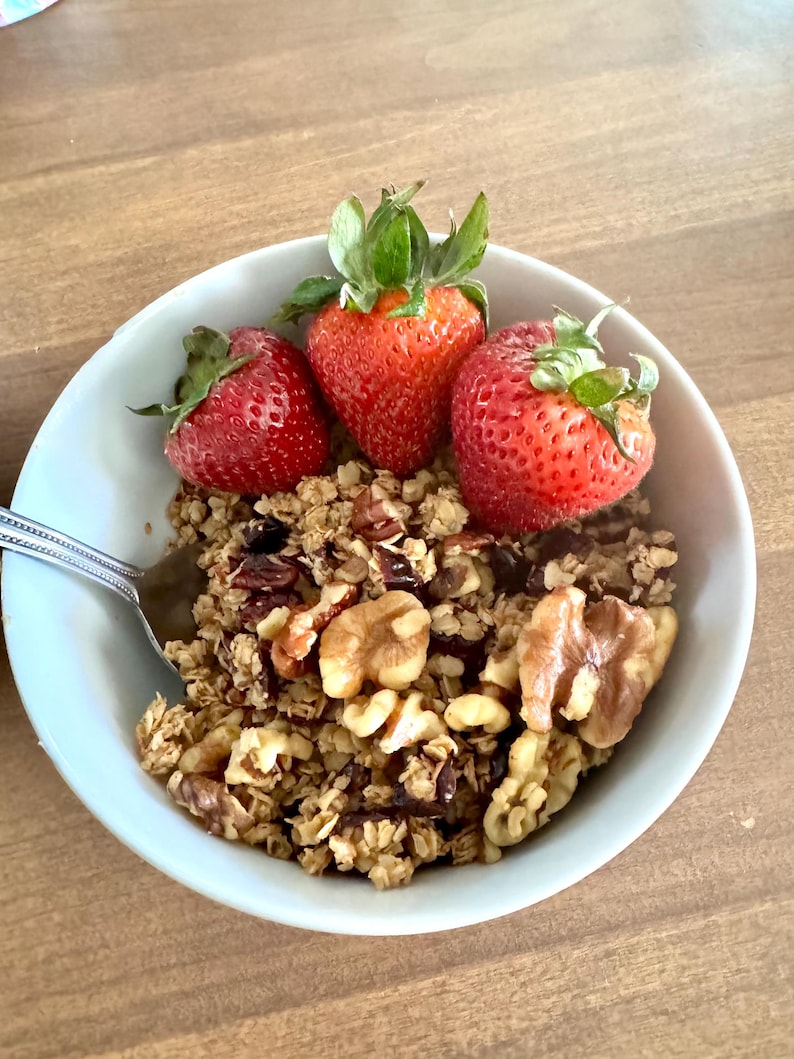 May include: A light blue bowl filled with granola, walnuts, and three fresh strawberries. A silver spoon rests in the bowl. The food is on a wooden table.