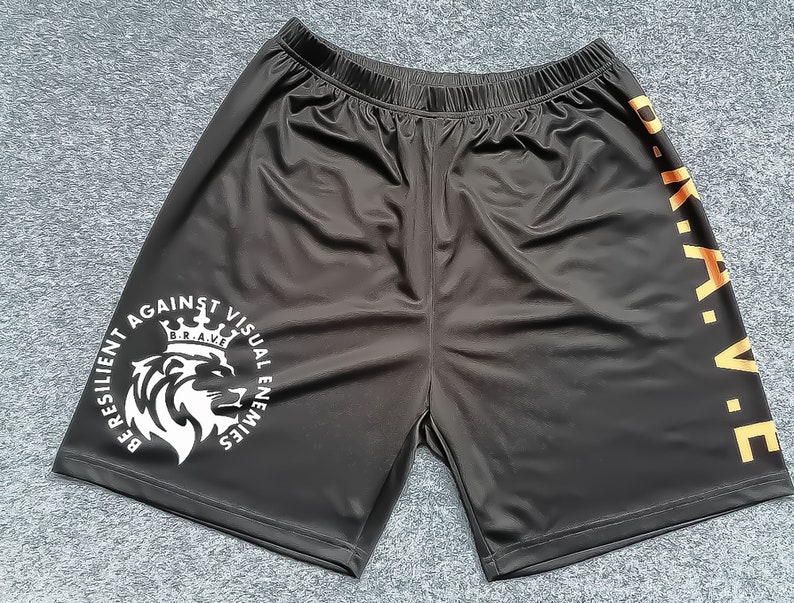 Op de afbeelding: Zwarte sport shorts met een witte leeuw afbeelding en de tekst "Be Resilient Against Visual Enemies" en "B.R.A.V.E." aan de zijkant.