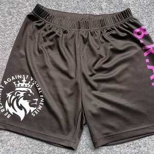 Op de afbeelding: Zwarte sport shorts met een witte afbeelding van een leeuw met een kroon en de tekst "BE RESILIENT AGAINST VISUAL ENEMIES" en de tekst "B.R.A.V.E." in roze aan de zijkant.