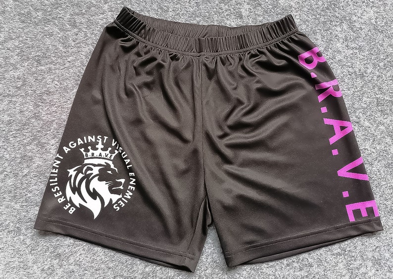 Op de afbeelding: Zwarte sport shorts met een witte afbeelding van een leeuw met een kroon en de tekst "BE RESILIENT AGAINST VISUAL ENEMIES" en de tekst "B.R.A.V.E." in roze aan de zijkant.