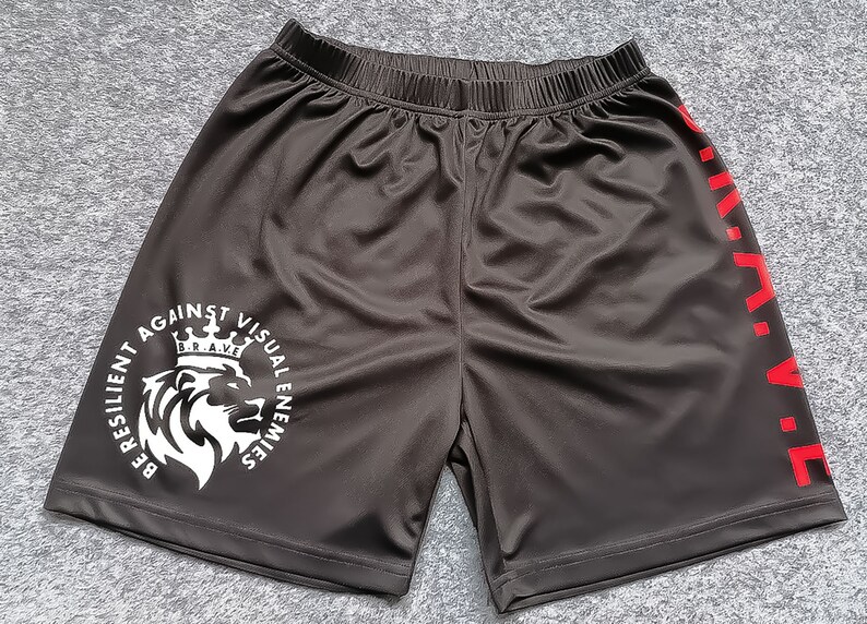 Op de afbeelding: Zwarte sport shorts met een witte leeuw afbeelding en de tekst "Be Resilient Against Visual Enemies" en een rode "B.R.A.V.E." aan de zijkant.