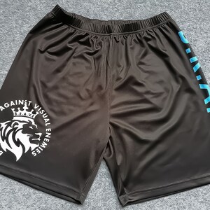 Op de afbeelding: Zwarte sport shorts met een witte afbeelding van een leeuw met een kroon en de tekst "BE RESILIENT AGAINST VISUAL ENEMIES B.R.A.V.E." Het rechterbeen heeft de tekst "B.R.A.V.E." verticaal in blauw gedrukt.
