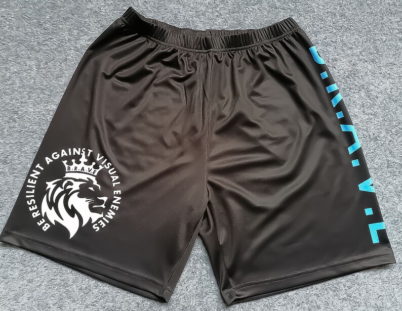 Op de afbeelding: Zwarte sport shorts met een witte afbeelding van een leeuw met een kroon en de tekst "BE RESILIENT AGAINST VISUAL ENEMIES B.R.A.V.E." Het rechterbeen heeft de tekst "B.R.A.V.E." verticaal in blauw gedrukt.