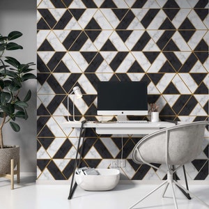 Op de afbeelding: Een wit bureau met een zwart metalen frame en een witte stoel. Het bureau heeft een zwart-wit geometrisch behang met gouden accenten. Een computermonitor, een lamp en een witte kom staan op het bureau.