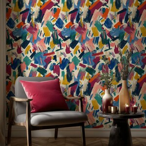 May include: A living room scene featuring a gray armchair with a red velvet pillow. The wall is covered in colorful abstract wallpaper with brushstroke patterns in blue, yellow, orange, and pink. A small table holds vases and candles.
