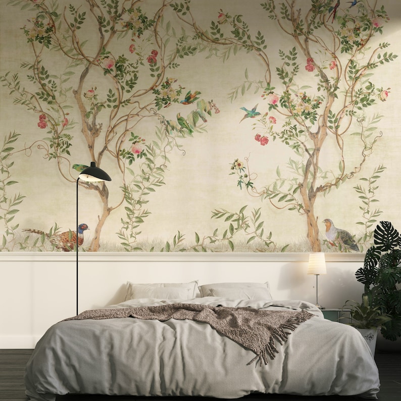 May include: A bedroom with a botanical mural featuring trees, birds, and flowers. The bed has gray bedding and a brown throw blanket. A black floor lamp and a small white bedside lamp provide lighting. A potted plant sits on the right.