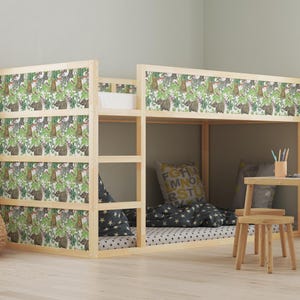 Puede incluir: Una litera de madera clara con cabecero y paneles laterales estampados con motivos de jungla. La cama tiene una litera inferior con ropa de cama y almohadas, y una pequeña mesa y taburete de madera están cerca. Una cesta tejida con animales de peluche está en primer plano.