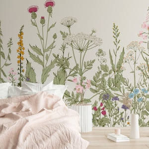 May include: A bedroom with a floral wallpaper featuring a variety of flowers and plants in shades of green, pink, yellow, and blue. A white bed with a pink blanket is in the foreground. A white vase with a flower arrangement is on the right side of the image.